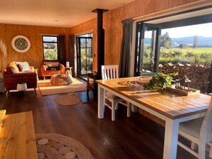 A cozy living room with a wooden floor, a dining table, and a sofa. There is a large window with a view of the outdoors.