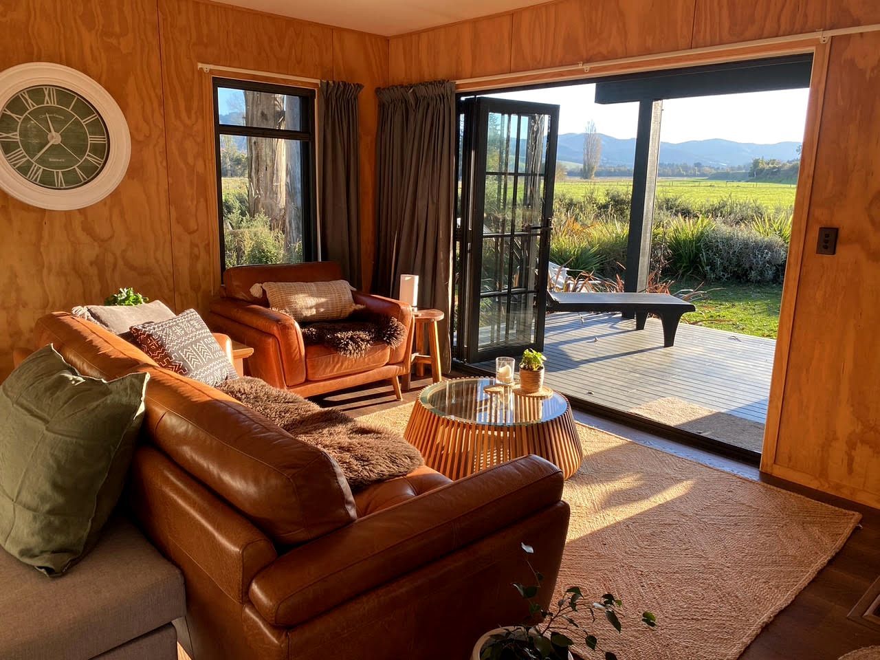 A room with a brown leather sofa, a glass coffee table, and a view of a deck and greenery outside.