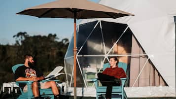 Two people sitting under a large umbrella in front of a tent.