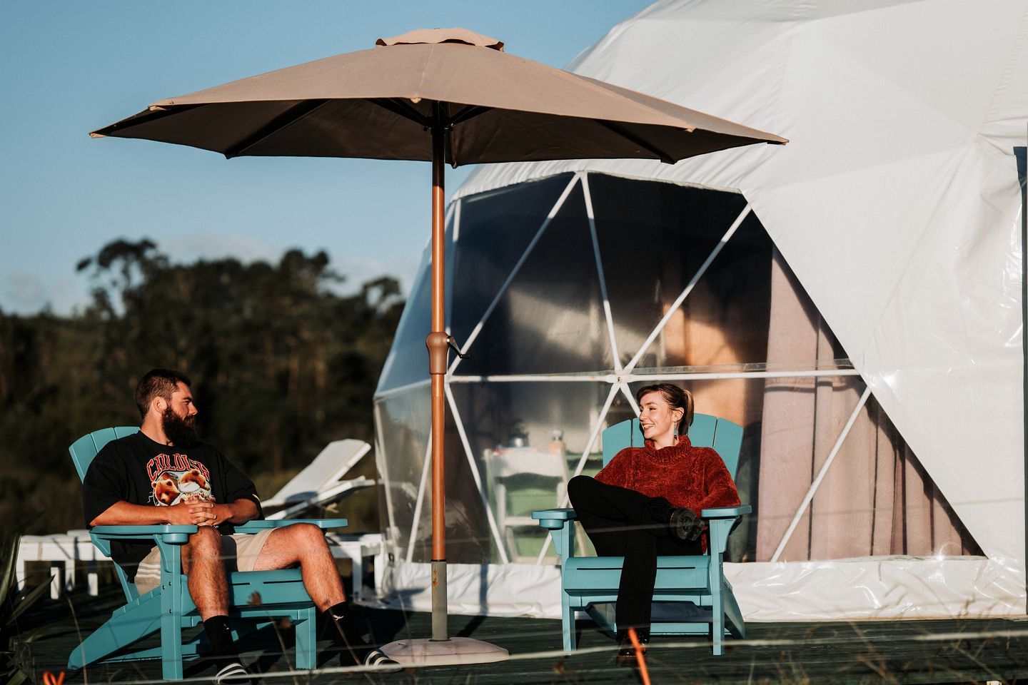 Two people sitting under a large umbrella in front of a tent.