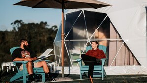 Two people sitting under a large umbrella in front of a tent.