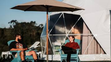 Two people sitting under a large umbrella in front of a tent.
