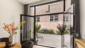 A modern room with a dining table, chairs, a vase with flowers, a potted plant, and a view of a brick building through a glass door. - Amstel l Family friendly l Fully equipped l Unit L (Amsterdam)