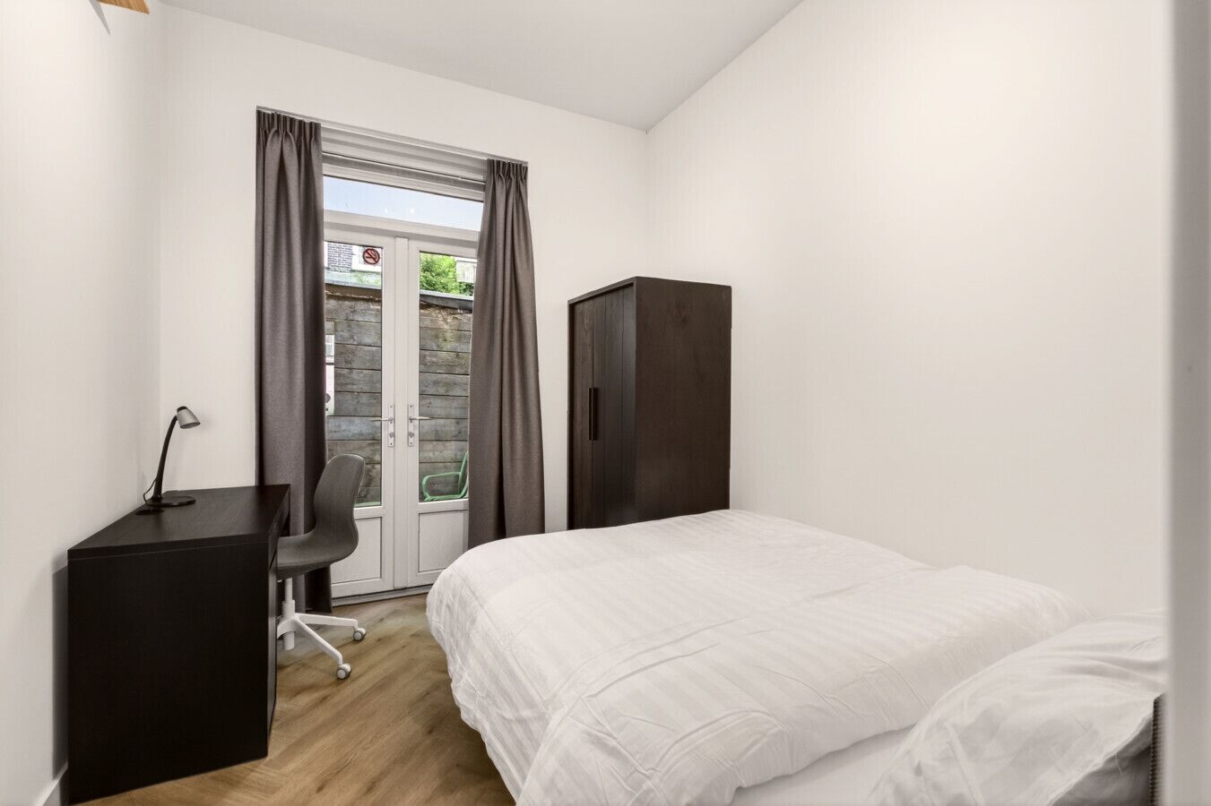 A bedroom with a bed, a desk, a chair, a wardrobe, and a sliding glass door leading to an outdoor area.