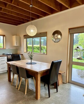 A dining area with a wooden table, chairs, a hanging light fixture, a clock, and a view of the outdoors through large windows. - Deluxe Cabin Rental in Stunning Setting near Tasman, South Island (Mahana)
