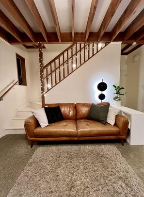 A brown leather sofa with pillows, a staircase with string lights, and a wall-mounted light fixture. - Deluxe Cabin Rental in Stunning Setting near Tasman, South Island (Mahana)