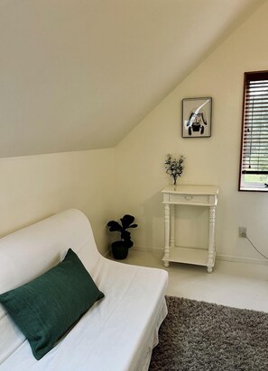 A room with a white sofa, a small white table with a plant, a framed picture on the wall, and a window with blinds. - Deluxe Cabin Rental in Stunning Setting near Tasman, South Island (Mahana)