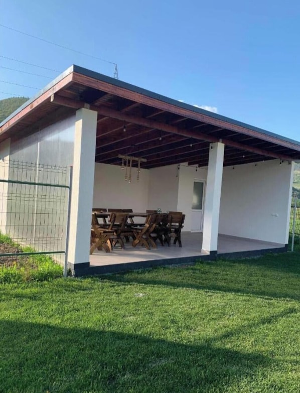 Ein überdachter Sitzbereich im Freien mit Tisch und Stühlen, einem eingezäunten Bereich und einem Rasen.