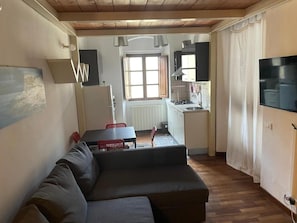A living room with a sofa, a dining table with chairs, a kitchen area with a stove and sink, and a television mounted on the wall. - Loft Terrazza in Acquaviva (Livorno)