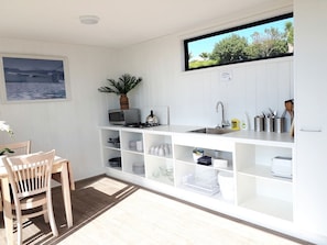 A compact kitchen with white cabinetry, a dining area with a wooden table and chairs, a potted plant, and a view of the outdoors.