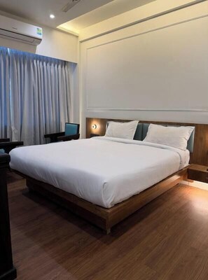 A hotel room with a large bed, a desk, a chair, a nightstand, and an air conditioning unit. - The Shivanta Inn (Abu Road)