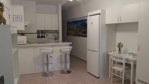 Cocina con armarios blancos, refrigerador, pileta y dos taburetes de bar, junto a una mesa pequeña con una planta.