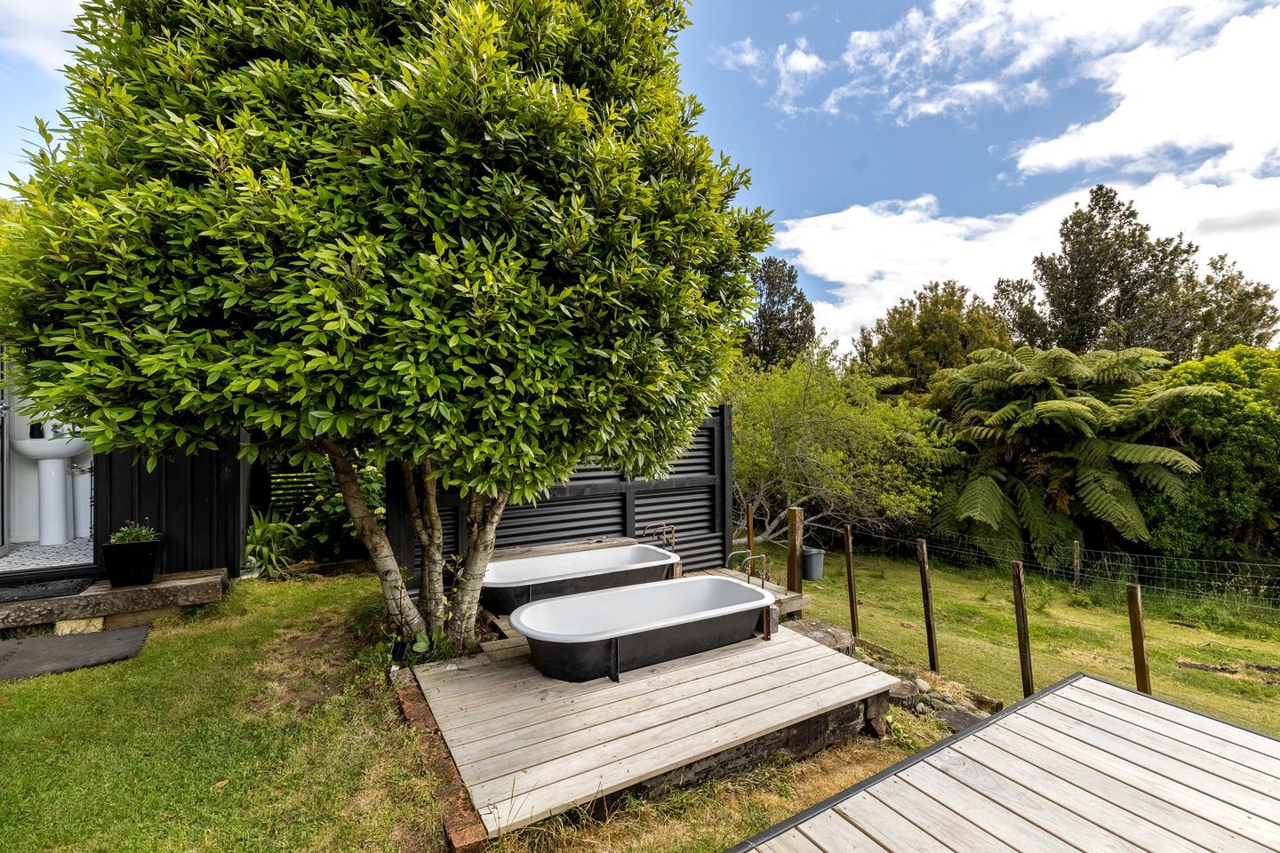 A backyard with a wooden deck, two black hot tubs, a large tree, and a fenced area with greenery.