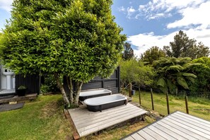 A backyard with a wooden deck, two black hot tubs, a large tree, and a fenced area with greenery.