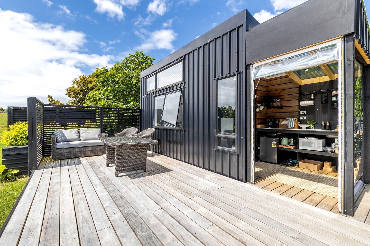 A modern outdoor deck with a sofa and table, a kitchen area with cabinets and appliances, and a large window.