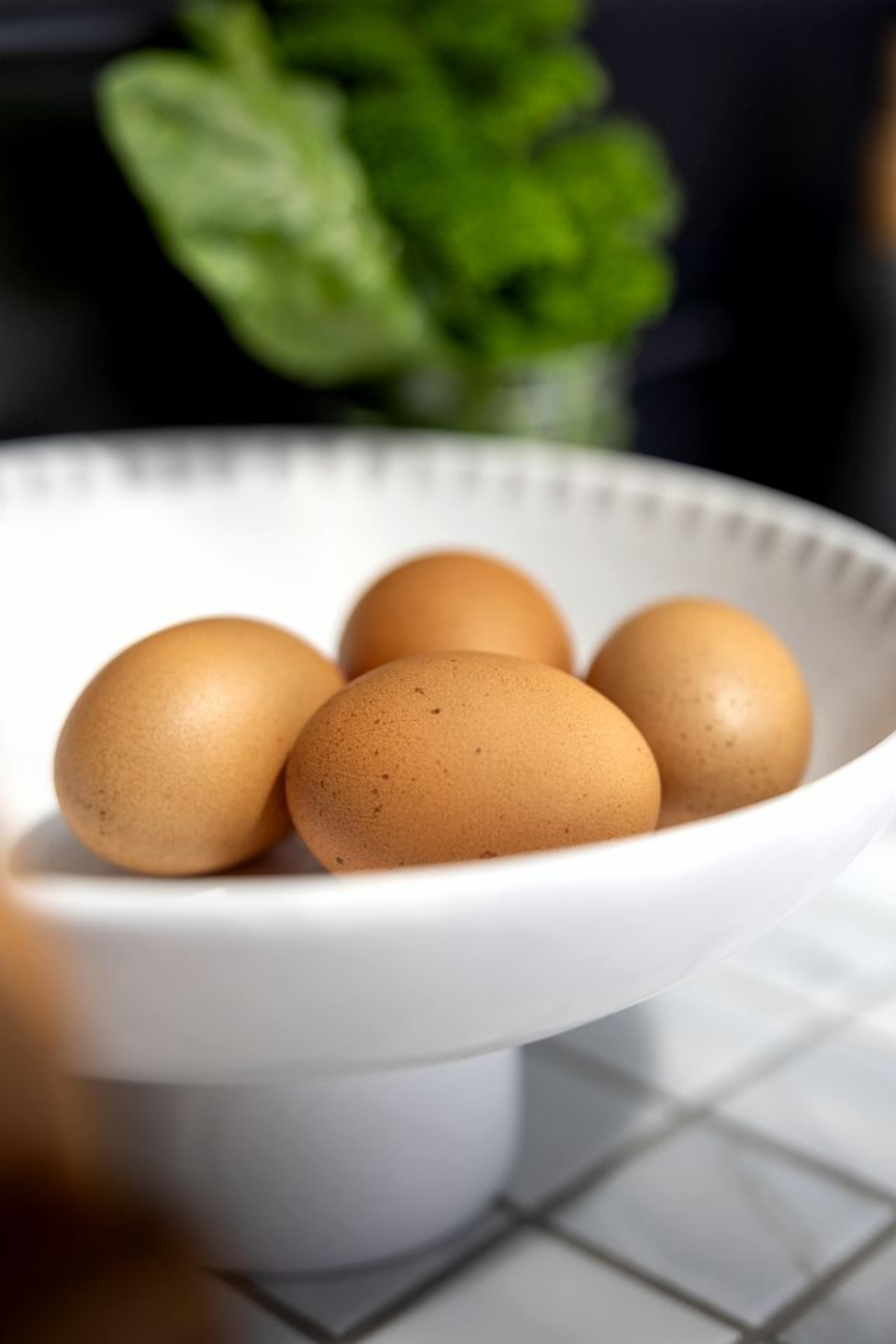 A white bowl containing five brown eggs on a white surface with a blurred green plant in the background.
