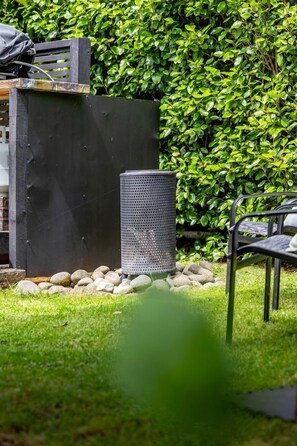 Ein Gartenbereich mit einem Metallstuhl, einer schwarzen Mülltonne und einer grünen Hecke im Hintergrund.