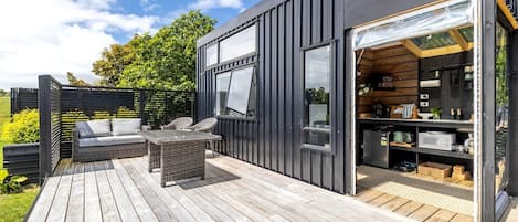 Une terrasse extérieure moderne avec un canapé et une table, un coin cuisine doté de placards et d’appareils électroménagers, et une grande fenêtre.