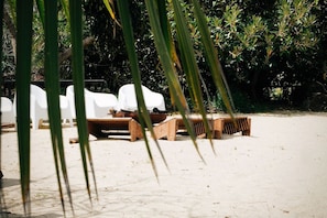 Un área de descanso frente a la playa con sillas de plástico blanco y bancos de madera, rodeada de abundante vegetación.