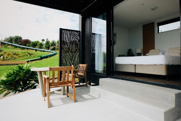 A modern outdoor seating area with a table and chairs, a bedroom with a bed and bedside table, and a view of a green landscape.