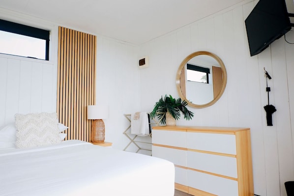 Un dormitorio con cama, mesita de noche, lámpara, espejo, televisor y una planta.