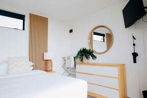 Un dormitorio con cama, mesita de noche, lámpara, espejo, televisor y una planta.