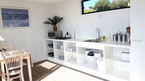 Eine kompakte Küche mit weißen Schränken, ein Essbereich mit Holztisch und -stühlen, eine Topfpflanze und Blick ins Freie.