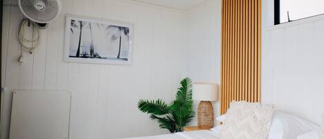 A bedroom with a bed, a wall-mounted fan, a framed picture, a potted plant, and a wooden slat wall panel.