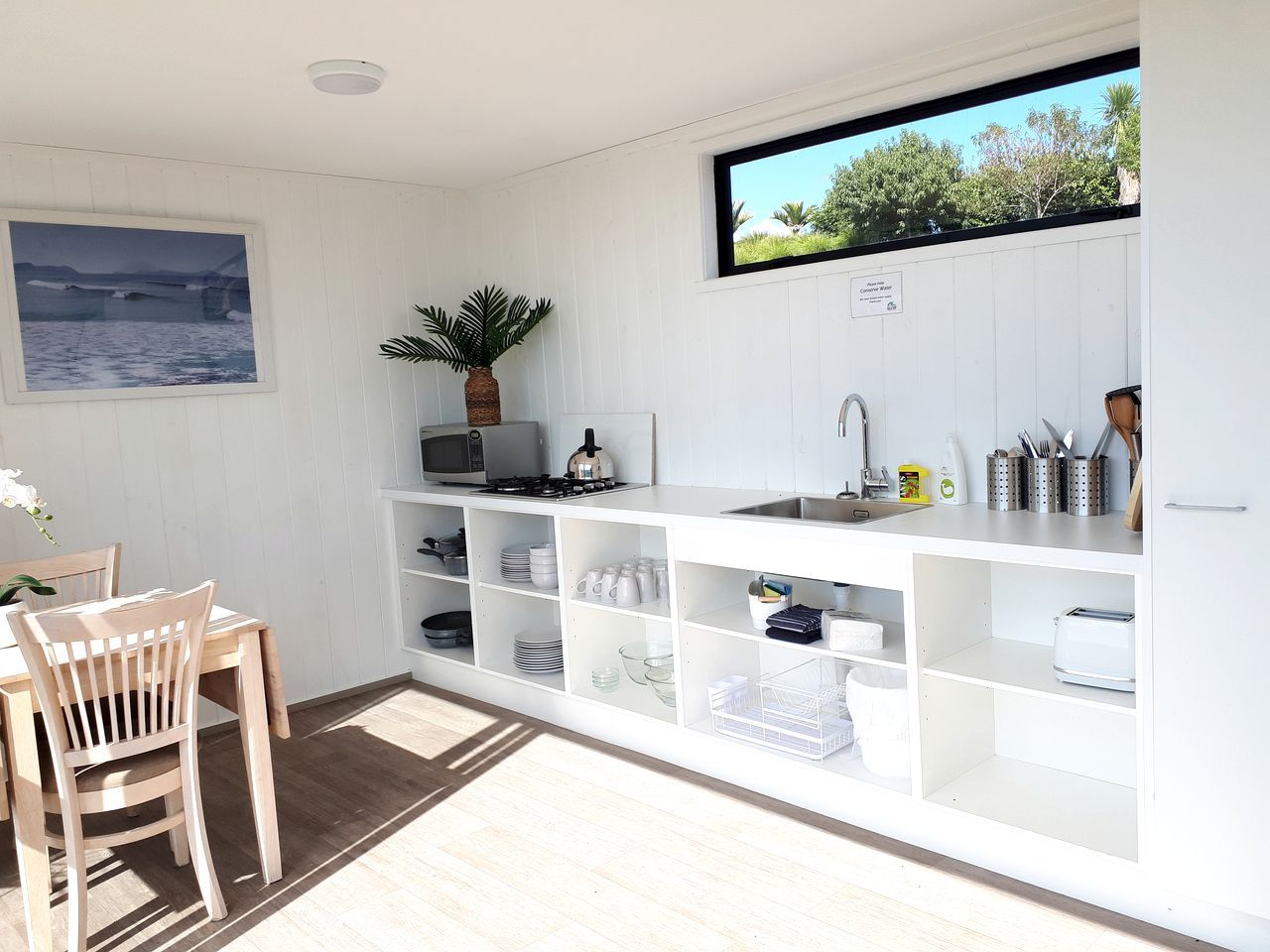 A compact kitchen with white cabinetry, a dining area with a wooden table and chairs, a potted plant, and a view of the outdoors.