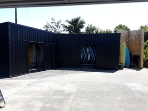 Ein überdachter Außenbereich mit Betonboden, einer schwarzen Metallkonstruktion und Surfbrettern, die an der Konstruktion angelehnt sind.