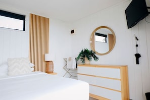 Ein Schlafzimmer mit Bett, Nachttisch, Lampe, Spiegel, Fernseher und einer Pflanze.