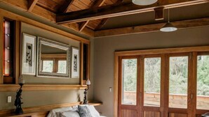 A bedroom with a wooden ceiling, a bed, a desk, and a window with a view of greenery.