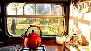 A kitchen with a red kettle on a stove, a window with a view of a garden, and a potted plant on the windowsill.