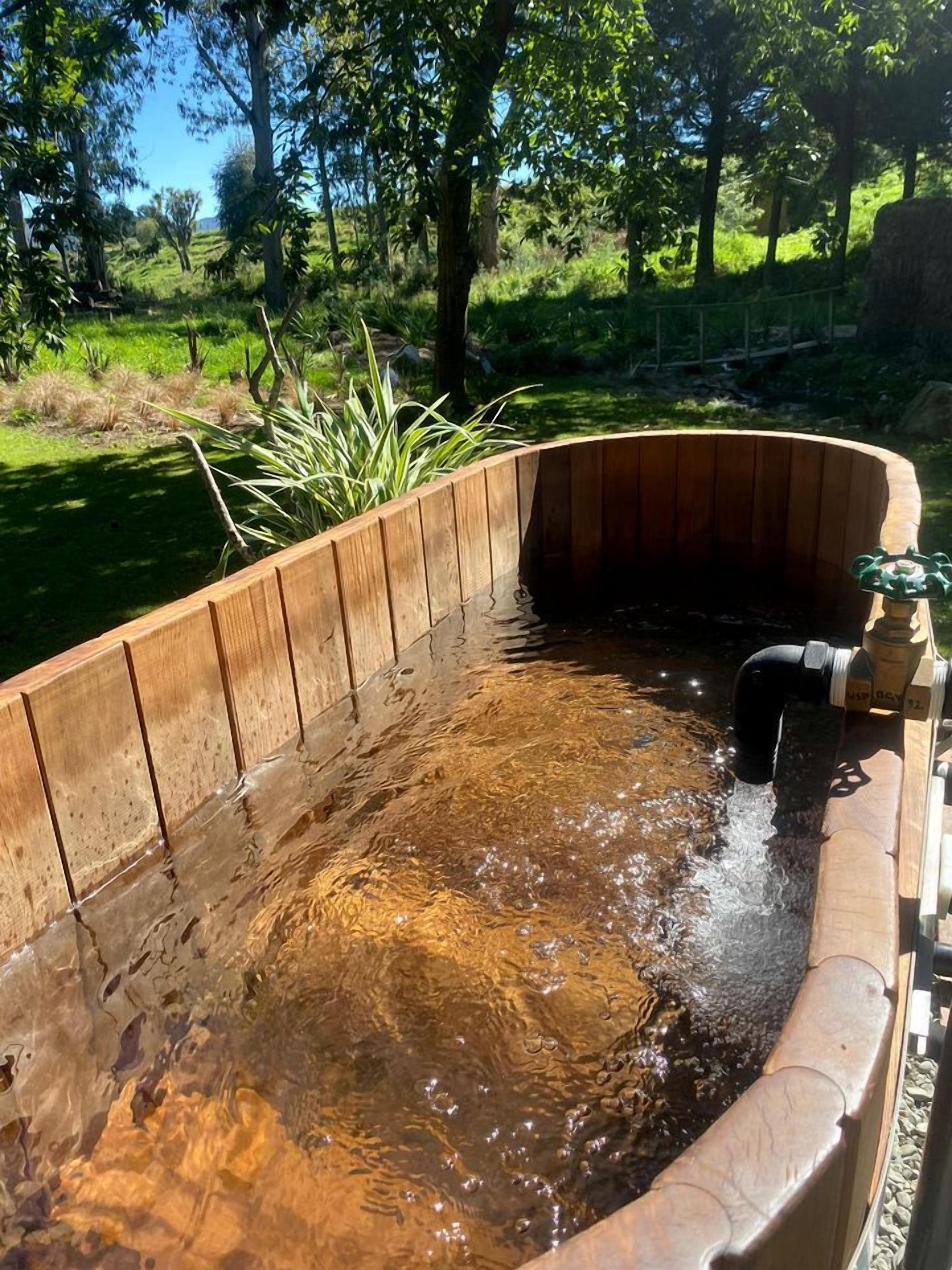 Un jacuzzi de madera con diseño curvo, rodeado de follaje y árboles.