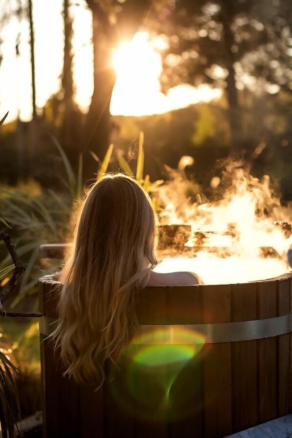 Una persona sentada en una bañera de madera al aire libre, con un fuego de fondo.