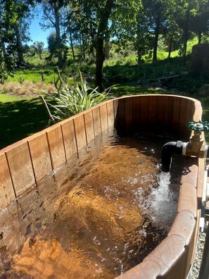 A wooden hot tub with a curved design, surrounded by greenery and trees.