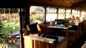 Een knus interieur met een houten bank, een tafel met verschillende spullen, en uitzicht op een tuin door een raam.