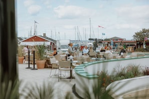 An outdoor seating area with tables and chairs, a red building, and sailboats in the background.