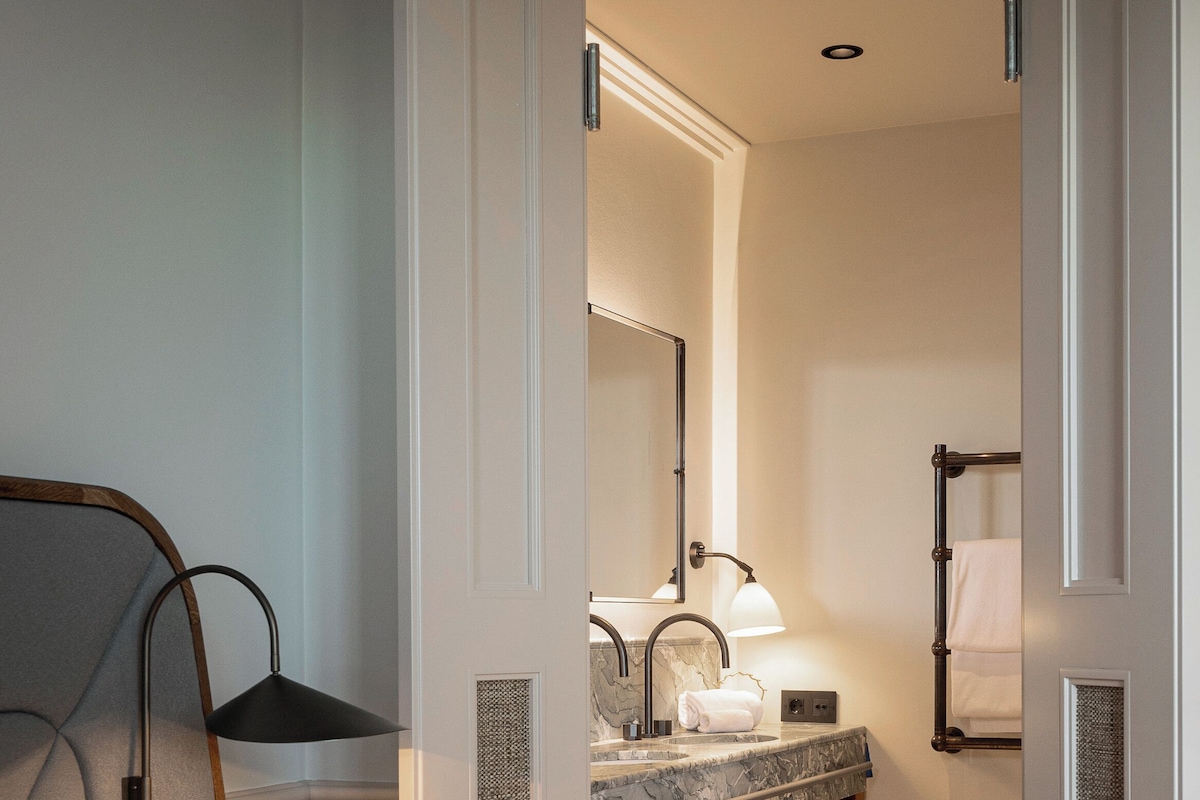 A hotel room with a bed, a bedside table, a wooden nightstand, a bathroom with a sink, mirror, towel rack, and a shower area.