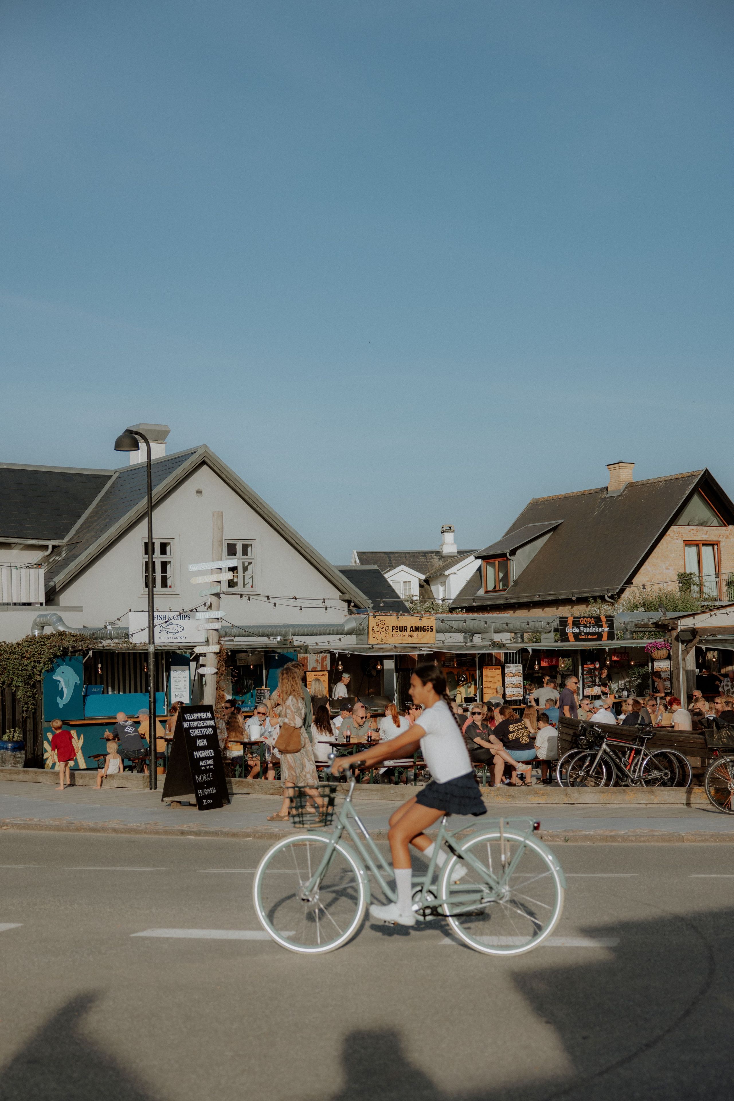 Une scène de rue où l’on voit un cycliste à vélo. On aperçoit des bâtiments avec des enseignes, un lampadaire et une foule de personnes.