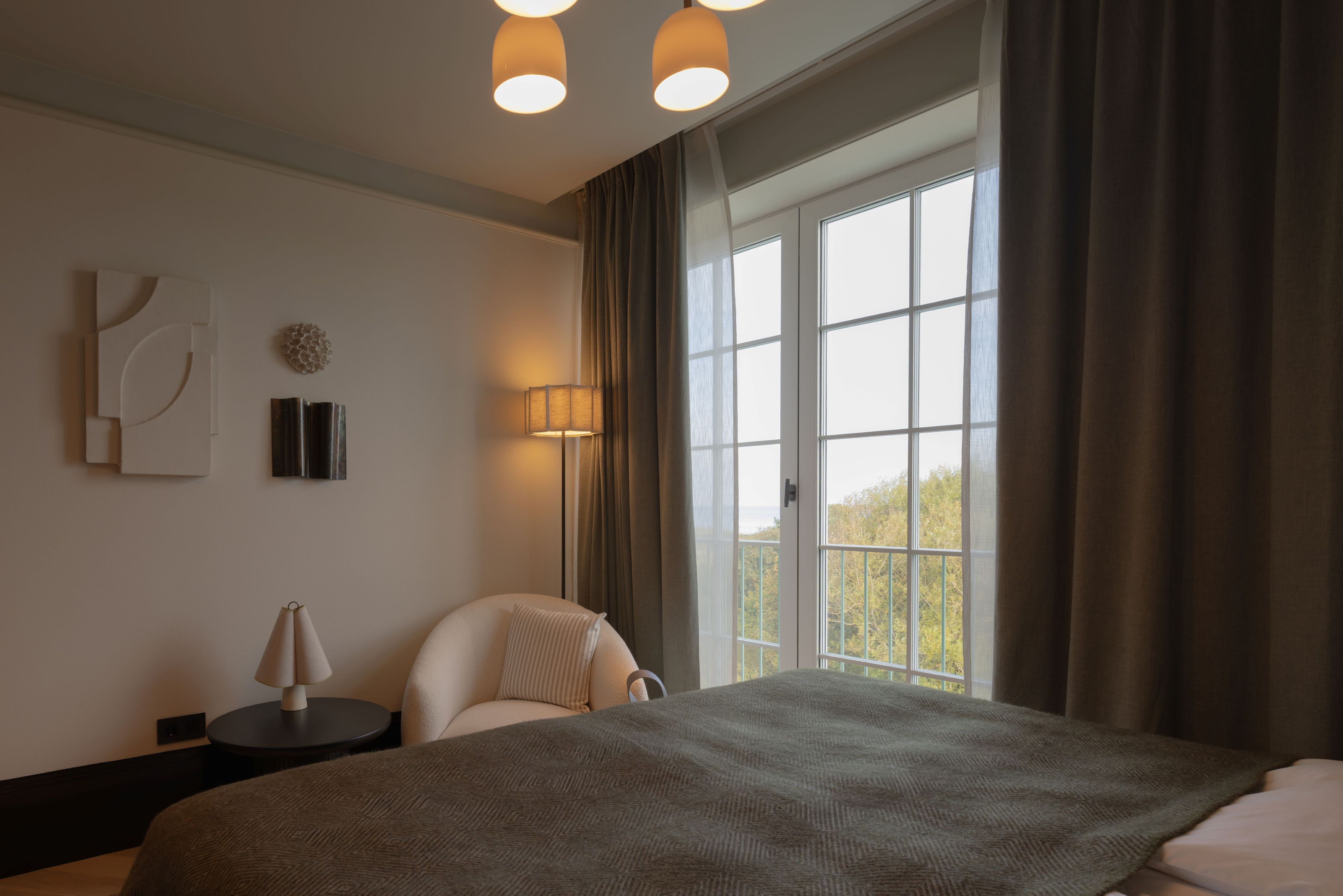 Habitación de hotel con una cama grande, una mesita de noche, una lámpara, una silla y vistas al exterior a través de una ventana.