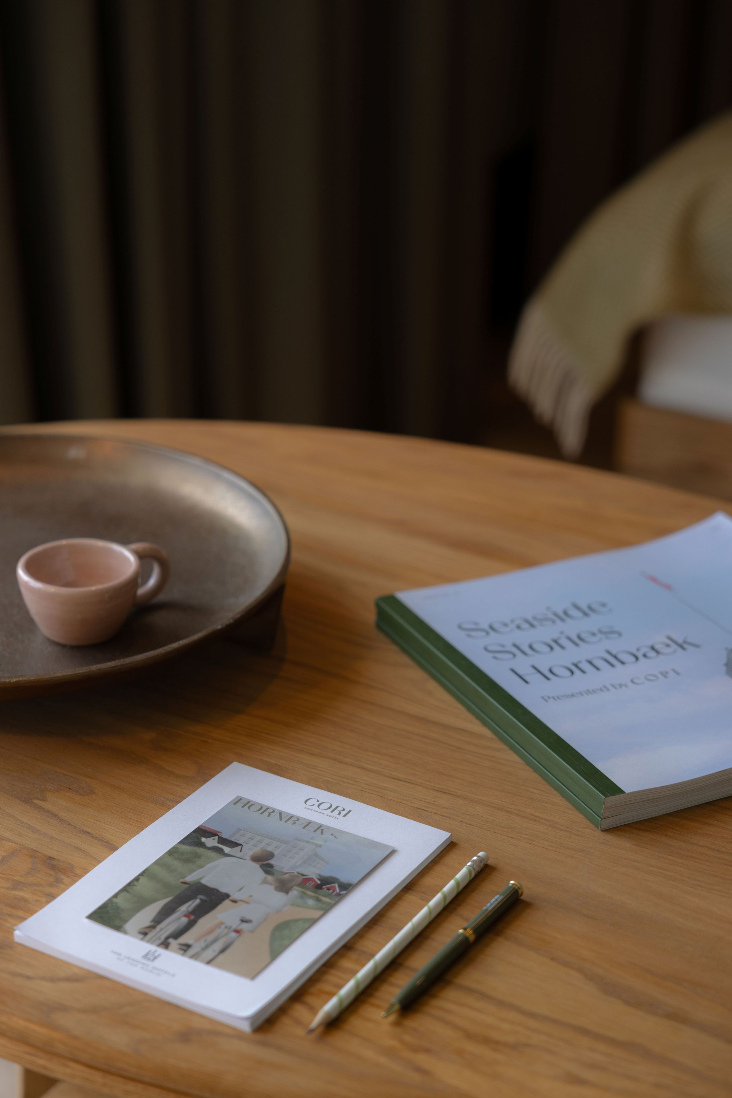 Une table en bois sur laquelle se trouve un livre intitulé « Seaside Slow Stories Hornback » et une revue dont la couverture représente un homme et une maison. Il y a deux stylos sur la table.
