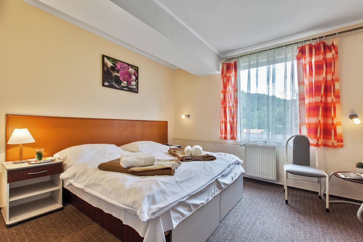 Ein Hotelzimmer mit Bett, Nachttisch, Lampe, Fenster mit Vorhängen, Stuhl und einem Bild an der Wand.