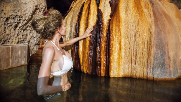 Una persona con traje de baño blanco tocando una gran formación rocosa y texturizada en un entorno similar a una cueva.