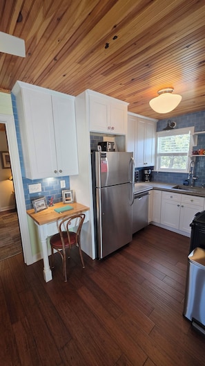 A compact kitchen with wooden flooring, white cabinets, a stainless steel refrigerator, and a wooden dining table with a chair.