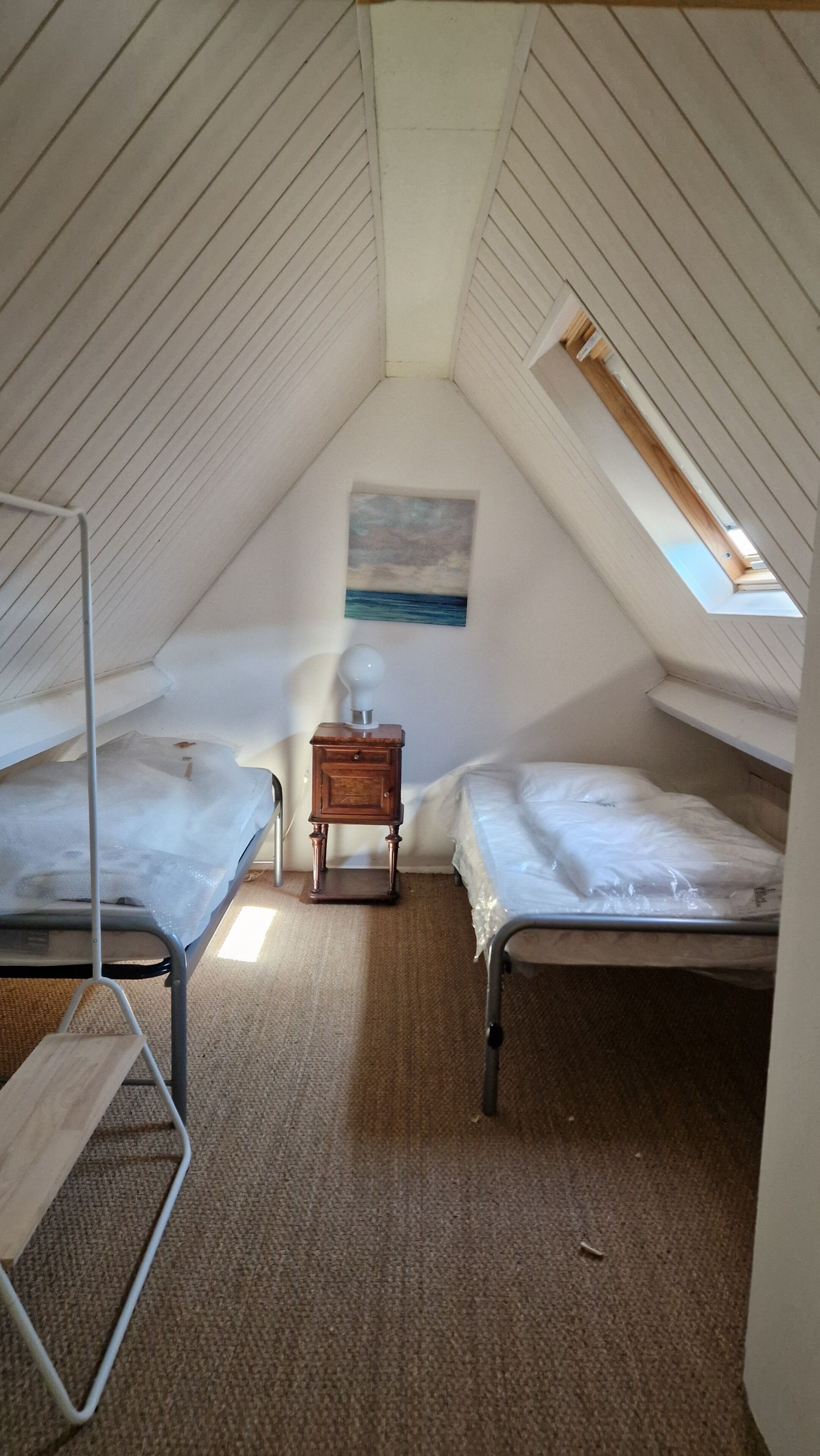 Ett litet sovrum på vinden med två våningssängar i metall, ett nattduksbord i trä, en enkel lampa och en tavla på väggen.