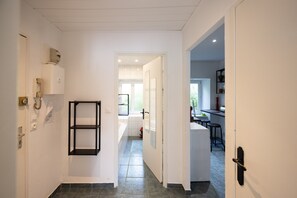 Ein Flur, der zu einem Badezimmer mit Toilette, Waschbecken und Dusche führt. Rechts befindet sich eine Tür zu einem Küchenbereich mit Arbeitsfläche und Hockern.