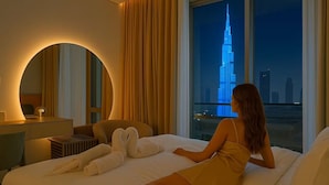 A hotel room with a bed, a round mirror, a desk with a lamp, and a view of a tall building.