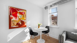 A room with a white wall, a large abstract painting, a round table with two chairs, a gray sofa, and a window with blinds.