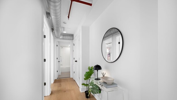 A hallway with white walls, a wooden floor, a round mirror, a black lamp, a white bowl, and a potted plant.
